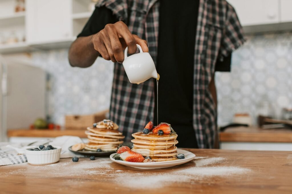 A person pouring maple syrup over a stack of pancakes topped with fresh berries.